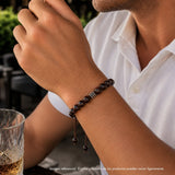 Modelo con pulsera tejida para hombre modelo Ares con hilo marrón, esferas de rodonita borgoña y tres aros de plata 925 quemada sobre fondo blanco.